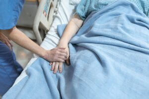 An unknown nurse holds the hand of an unknown patient. The patient is in a hospital bed with blankets.