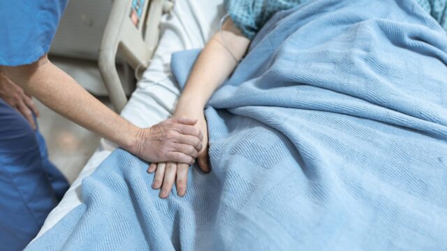 An unknown nurse holds the hand of an unknown patient. The patient is in a hospital bed with blankets.