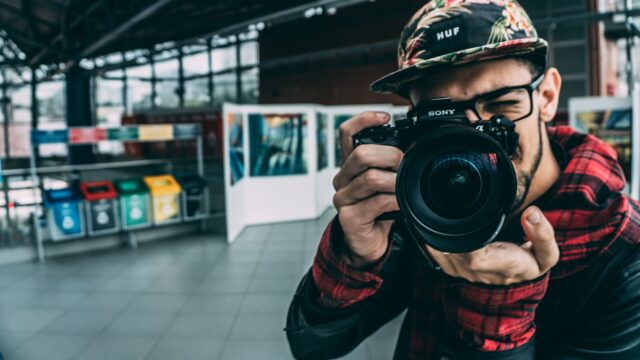 A person points a camera towards the viewer. There are displays of photography in the background.