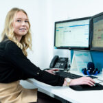 An apprentice works at a desk that has a computer, plant pot and phone on it.