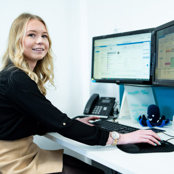 An apprentice works at a desk that has a computer, plant pot and phone on it.