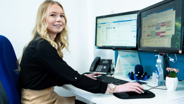 An apprentice works at a desk that has a computer, plant pot and phone on it.
