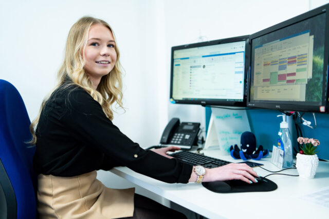 An apprentice works at a desk that has a computer, plant pot and phone on it.