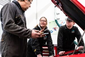 People in overalls gather under the bonnet of a car.