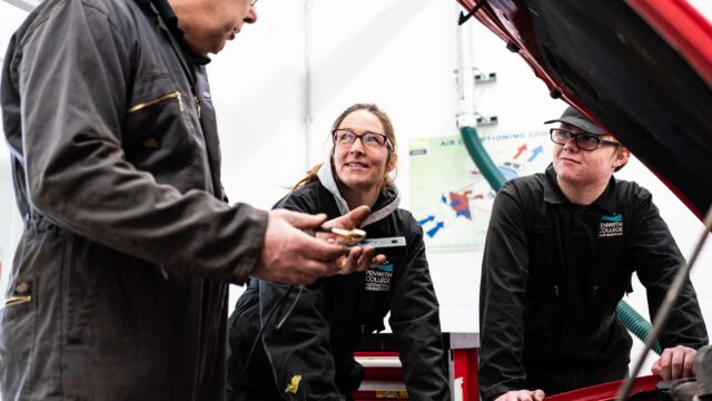 People in overalls gather under the bonnet of a car.