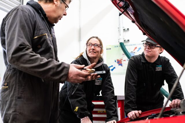 People in overalls gather under the bonnet of a car.