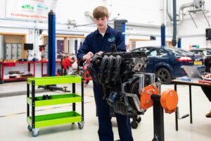 A student in blue overalls is working on a car engine.