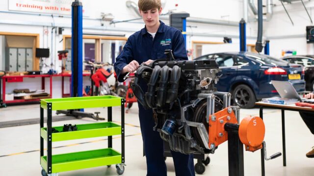 A student in blue overalls is working on a car engine.