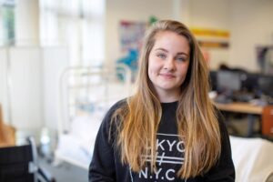 A close up of a student with long hair and wearing a dark jumper. There is a hospital bed in the background.
