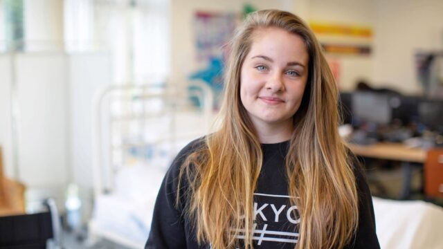 A close up of a student with long hair and wearing a dark jumper. There is a hospital bed in the background.