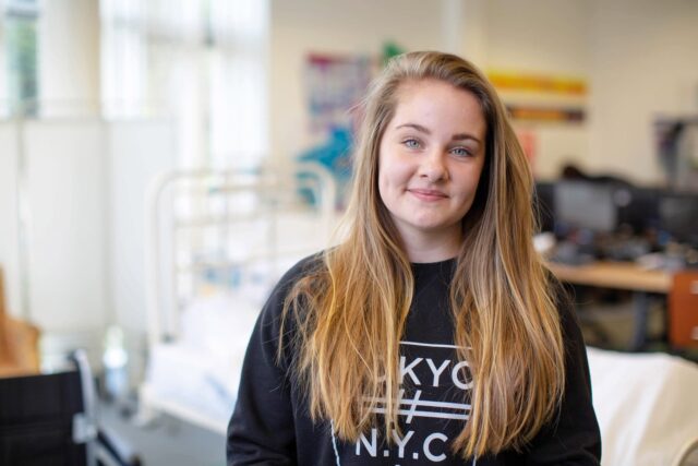 A close up of a student with long hair and wearing a dark jumper. There is a hospital bed in the background.