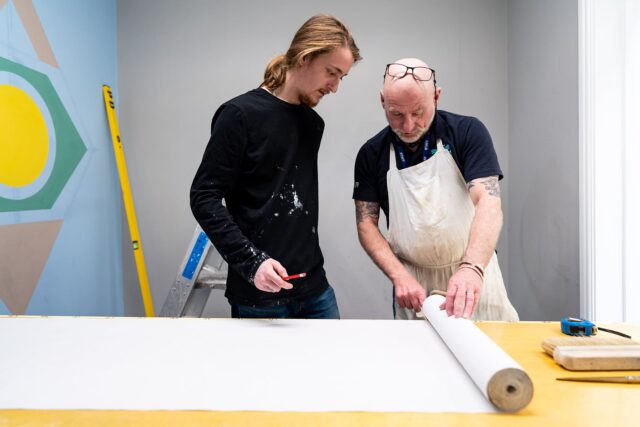 A tutor and a student look at a roll of wallpaper together.