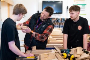 Two students look to a tutor in a workshop. The tutor holds up a piece of wood.
