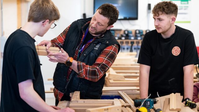 Two students look to a tutor in a workshop. The tutor holds up a piece of wood.