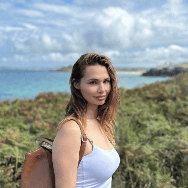 Bethany faces the camera with the sea in the background. She has a rucksack on one shoulder.