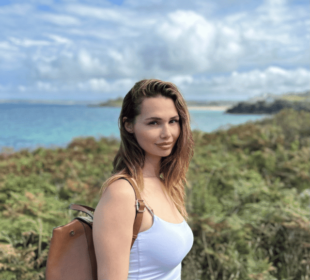Bethany faces the camera with the sea in the background. She has a rucksack on one shoulder.