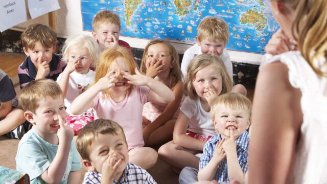 Children sit on the floor facing an unrecognisable teacher and all point towards their noses.