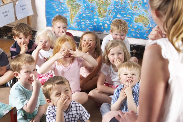 Children sit on the floor facing an unrecognisable teacher and all point towards their noses.