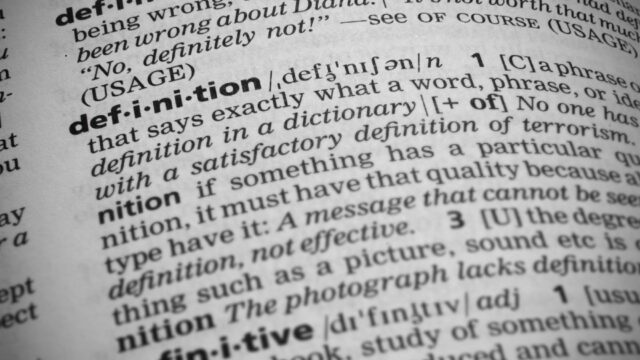 Close-up shot of the word definition in a dictionary. Page curves and has a vignetting or edge burn effect where the corners are darker. Focus is on the word definition.
