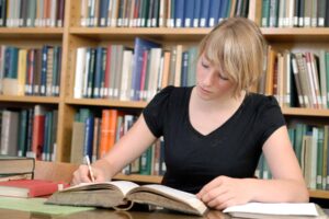 A student sits in a library making notes with a book open in front of her.