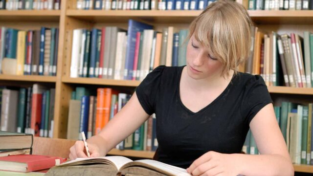 A student sits in a library making notes with a book open in front of her.