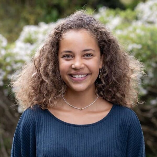 Student, Kaeya, stands outside with her hands behind her back. She has curly hair and is wearing a necklace, blue jumper and white trousers.