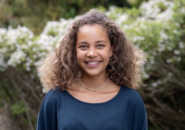 Student, Kaeya, stands outside with her hands behind her back. She has curly hair and is wearing a necklace, blue jumper and white trousers.