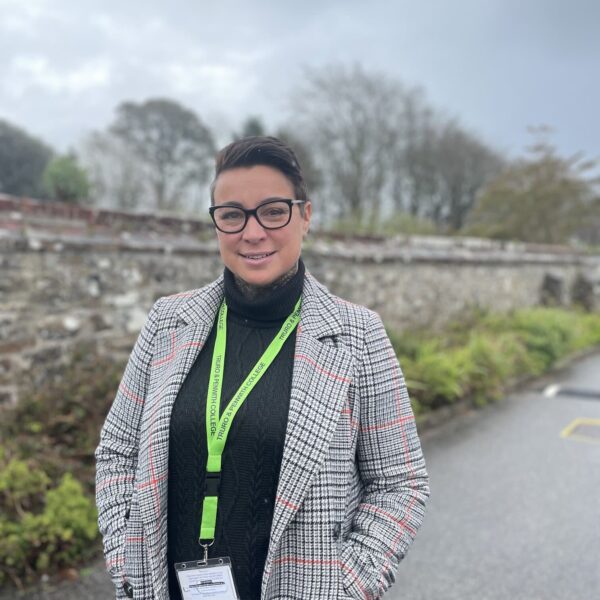 A photo of Kelly stood outside with her hands in her coat pockets. She is wearing a green lanyard and black galsses.