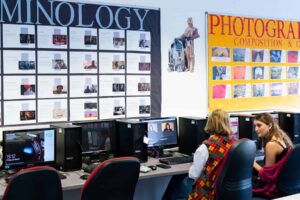 An image of two students sat in a computer room. There are posters on the wall in front of them.