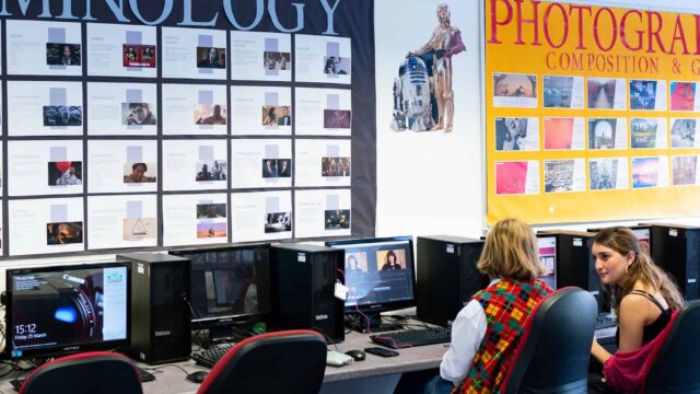 An image of two students sat in a computer room. There are posters on the wall in front of them.