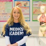 A student with long blonde hair looks at the camera. She is wearing a blue T-shirt that says 'Medics' Academy' on it. There is a dummy of a human body in the background.