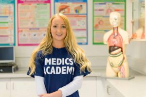 A student with long blonde hair looks at the camera. She is wearing a blue T-shirt that says 'Medics' Academy' on it. There is a dummy of a human body in the background.