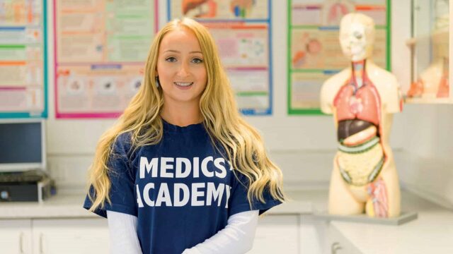 A student with long blonde hair looks at the camera. She is wearing a blue T-shirt that says 'Medics' Academy' on it. There is a dummy of a human body in the background.