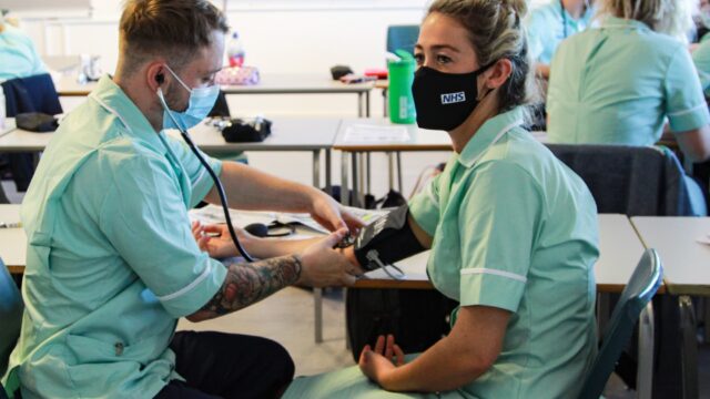 A student uses a stethoscope on another student. They are both wearing nurse uniforms.