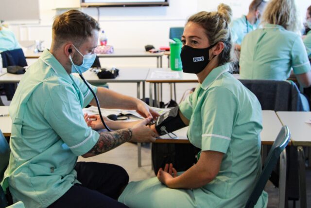 A student uses a stethoscope on another student. They are both wearing nurse uniforms.