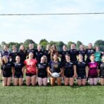 The female rugby academy in a group photo on a rugby pitch underneath the goal posts.