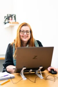A girl with red hair sits in front of a laptop and smiles at the camera.