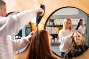 A photo of a hairdresser blow drying the hair of a client. There are shampoo and conditioner bottles in the background.