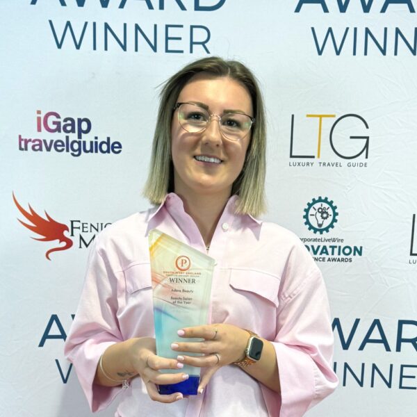 A photo of a woman holding up a trophy. She is wearing a pink shirt and is stood against a banner with logos on it.