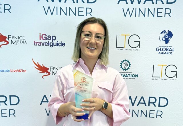 A photo of a woman holding up a trophy. She is wearing a pink shirt and is stood against a banner with logos on it.