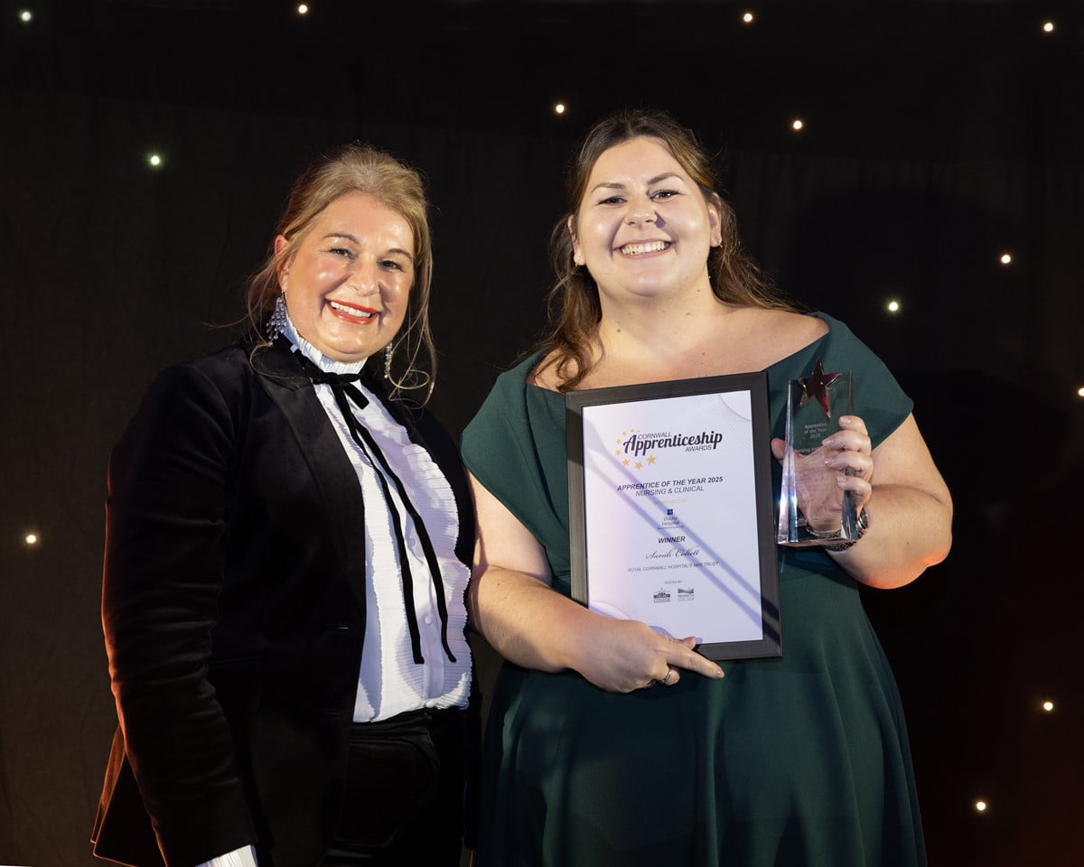 A student in a green dress holds up a certificate and a trophy and smiles. Another person stands with her in a black jacket and trousers and white shirt.