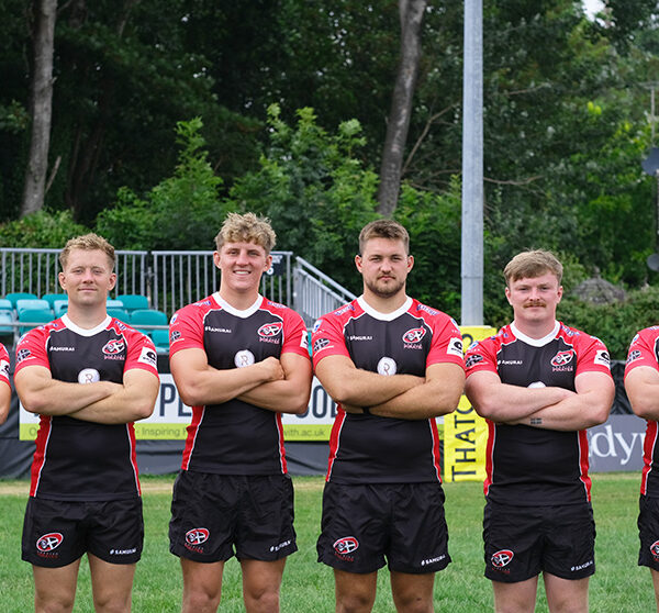 Cornish pirate rugby players stand in a line arms folded looking at the camera. They are stood on a pitch.