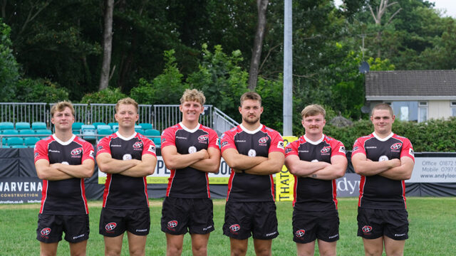 Cornish pirate rugby players stand in a line arms folded looking at the camera. They are stood on a pitch.