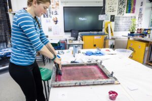 A student is printmaking in an art classroom. They are wearing a blue and white stripe shirt.