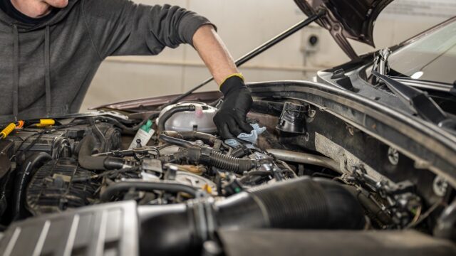 Mechanic In Gloves Servicing A Car Engine With Tools And Cloth In A Well Lit Garage.