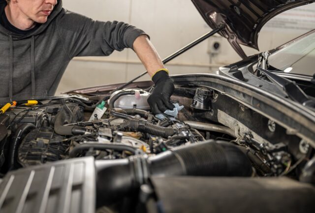 Mechanic In Gloves Servicing A Car Engine With Tools And Cloth In A Well Lit Garage.
