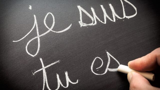 A hand writes out je suis tu es with chalk on a blackboard.