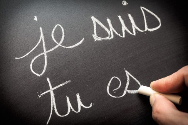 A hand writes out je suis tu es with chalk on a blackboard.