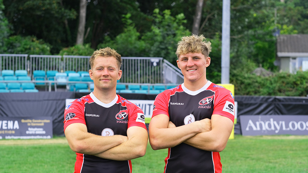 Two rugby players for Cornish Pirates fold their arms and look at the camera. They are stood on a rugby pitch.