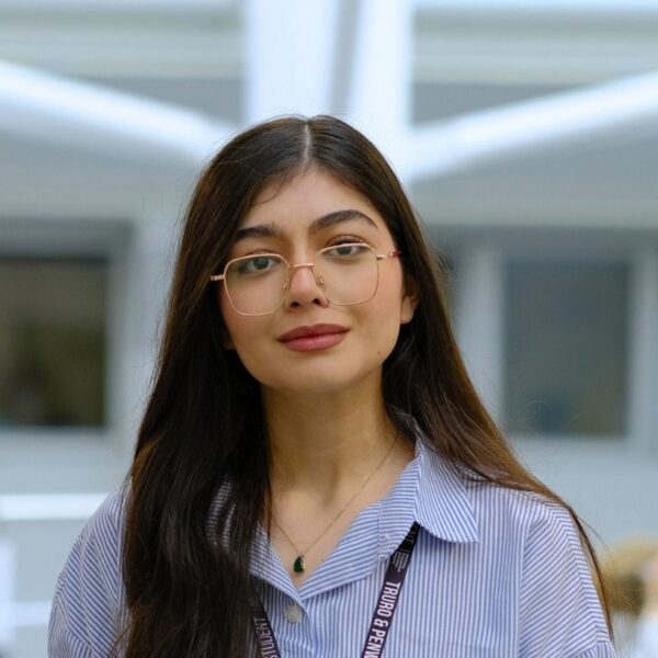 A student, Paria, stands looking at the camera. She wears a blue and white stripe shirt and has gold glasses.